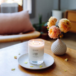 Brooklyn Candle Studio scented candle Kyoto series Escapist Mood photo of table with flowers. A romantic homage to Kyoto in the rain, green avenues and Japanese wooden architecture. This wonderful scented candle is a meditative, calming composition of Hinoki cypress, cedar, sandalwood, vetiver and pine. Hand-poured with 100% soy wax in the small manufactory of Brooklyn Candle Studio.